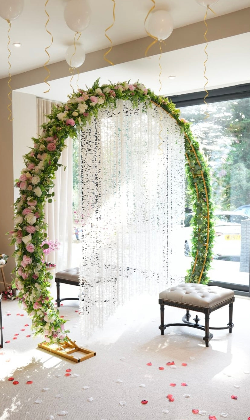 Decorative arch with flowers and hanging ribbons in a room with a bench and window.